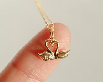 May include: A gold-colored necklace featuring a pendant with two swans forming a heart shape. The swans have a small diamond accent. The necklace is displayed on a finger against a neutral background. The text "emma's gems" is in the top right corner.