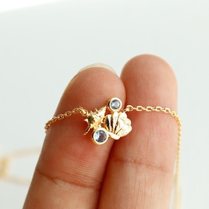 May include: A delicate gold necklace featuring seashell and starfish charms, accented with small blue gemstones. The necklace is held between fingers, showcasing the intricate details of the jewelry. The text "emma's gems" is visible.