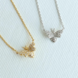 May include: Two delicate necklaces with bee pendants. One necklace is gold-toned, the other is silver-toned. Each bee pendant is encrusted with small, sparkling stones. The chains are thin and elegant, perfect for everyday wear.