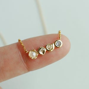 May include: A delicate gold necklace featuring a row of charms: a pearl, a seashell, a spiral shell, and a clear gemstone. The necklace is displayed on a finger against a soft, neutral background. The text "emma's gems" is in the upper right corner.