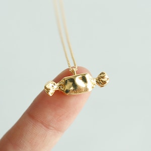 May include: A gold-colored candy pendant necklace. The pendant is shaped like a wrapped candy with textured details. The necklace is on a thin gold chain. The text "emma's gems" is in the top right corner.