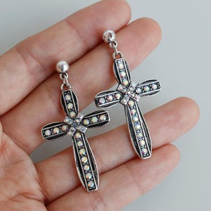 May include: A pair of silver-toned cross earrings with a dangling design. The crosses are embellished with iridescent crystals and small, round accents. The earrings are held in a hand.