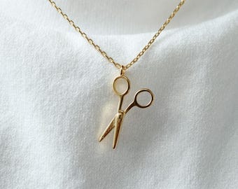 May include: A gold-colored necklace with a small scissors pendant. The scissors are detailed and hang from a delicate chain. The necklace is displayed against a white background. The text "emma's gems" is visible.
