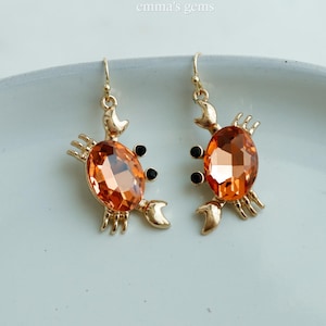 May include: A pair of gold-toned crab earrings. Each earring features an orange, oval-shaped gemstone body, black eyes, and crab claw details. The earrings are displayed on a white surface, with the text "emma's gems" in the top center.
