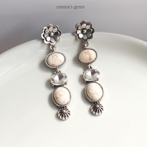 May include: A pair of silver-toned dangle earrings. Each earring features a floral top, oval cream-colored stones, clear crystals, and a floral bottom. The earrings are displayed on a white surface. The text "emma's gems" is in the top center.