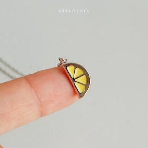 May include: A silver necklace with a pendant shaped like a lemon slice. The pendant has a yellow enamel filling and is held by a finger. The text "emma's gems" is in the upper right corner.
