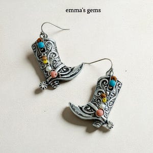 May include: Silver-toned cowboy boot earrings with intricate scrollwork and colorful bead accents. The earrings feature turquoise, yellow, pink, and white beads, and a small spur detail. The text "emma's gems" is visible.