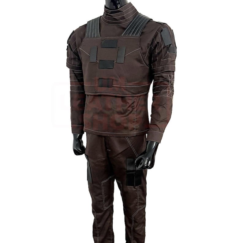 May include: A brown tactical jumpsuit with a high collar, long sleeves, and matching pants. The outfit features black accents on the chest, shoulders, and pockets, with visible stitching details. The mannequin is wearing black gloves.