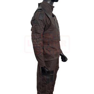 May include: Brown tactical uniform with black accents, displayed on a mannequin. The outfit features a high collar, shoulder pads, and multiple pockets. White stitching details the seams and geometric patterns on the sleeves. The uniform is designed for functionality and durability.