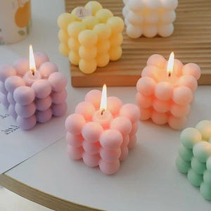 以下が含まれることがあります： カラフルなバブルキューブキャンドルのコレクション。キャンドルはピンク、紫、黄色、白、緑の色合いです。各キャンドルには火のついた芯があり、温かい光を放っています。キャンドルは白い表面と木製の板の上に配置されています。