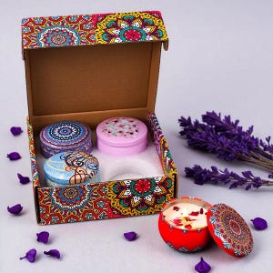 Könnte beinhalten: Ein Geschenkset mit vier dekorativen Kerzen in runden Dosen, präsentiert in einer gemusterten Box. Die Dosen weisen verschiedene Designs auf, darunter Blumen- und Mandala-Muster. Eine rote Kerze ist geöffnet und zeigt einen brennenden Docht und Rosenblätter.