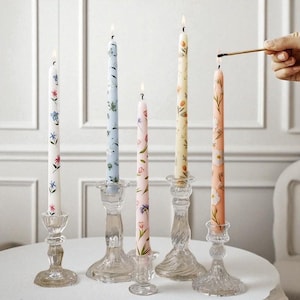 Może przedstawiać: Pięć kwiatowych świec stożkowych w szklanych świecznikach. Świece są białe, jasnoniebieskie, różowe i brzoskwiniowe, każda z innym kwiatowym wzorem. Ręka zapala świecę zapałką.