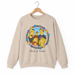 Op de afbeelding: Beige sweatshirt met een kleurrijke cirkelvormige afbeelding met een cartoonfamilieportret. De afbeelding bevat de tekst "Mom's Crew" in een handschriftstijl. De sweatshirt hangt aan een houten hanger.