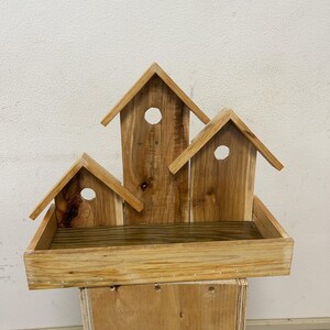May include: A wooden birdhouse with three individual houses and a rectangular tray. Each house has a circular opening and a pitched roof. The birdhouse is made of light-colored wood and is mounted on a wooden box.