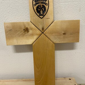 Könnte beinhalten: Holzkreuz mit einem Sheriff-Abzeichen-Design. Das Kreuz besteht aus hellfarbenem Holz, wobei das Sheriff-Abzeichen in den oberen Abschnitt geätzt ist. Das Abzeichen zeigt einen Stern und das Wort "SHERIFF".