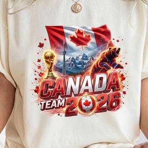 May include: Cream-colored t-shirt featuring a graphic design for the Canada Team 2026. The design includes the Canadian flag, a bear, the CN Tower, a soccer ball, and the FIFA World Cup trophy.