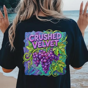 Peut inclure: T-shirt noir avec un motif coloré de raisins violets et de feuilles vertes, avec le texte "CRUSHED VELVET" en violet et vert. Le design est posé sur un fond bleu et violet.