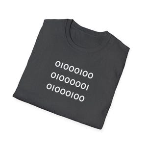 May include: Dark gray t-shirt with white binary code text. The text includes the numbers 0 and 1 in three rows. The shirt is folded and appears to be made of a soft material. The t-shirt has a crew neck.