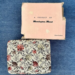 Pu&ograve; includere: Un astuccio vintage a fantasia floreale con una scatola rosa abbinata. L'astuccio presenta uno sfondo color crema con fiori rossi, gialli e marroni. La scatola recita "A Product of Remington Rand" e "(Patent Pending)".