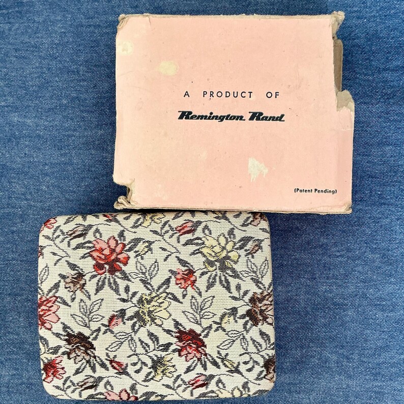 Pu&ograve; includere: Un astuccio vintage a fantasia floreale con una scatola rosa abbinata. L'astuccio presenta uno sfondo color crema con fiori rossi, gialli e marroni. La scatola recita "A Product of Remington Rand" e "(Patent Pending)".