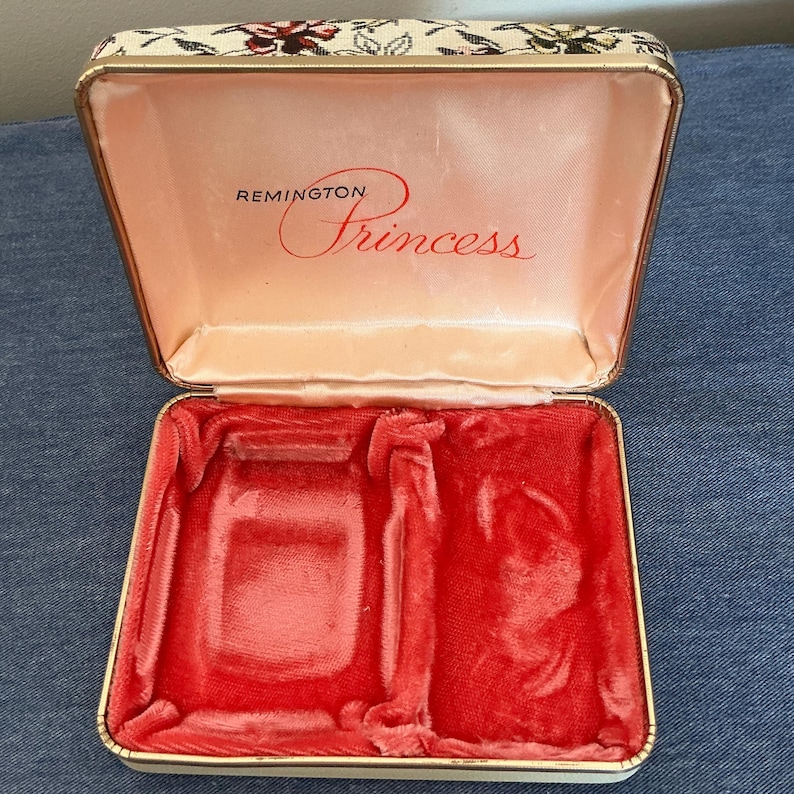 Pu&ograve; includere: Scatola vintage Remington Princess aperta. L'interno &egrave; foderato in raso rosa e velluto rosso. Il coperchio &egrave; decorato con un motivo floreale. La scatola &egrave; aperta, rivelando i vani interni.
