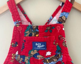 May include: Red corduroy baby overalls with a colorful print featuring cartoon characters and outdoor scenes. The overalls have adjustable straps, a front pocket with a "Baby B'gosh" label, and red buttons. The garment is hanging on a wooden hanger.