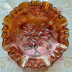 May include: An iridescent orange glass bowl with a ruffled edge. The bowl features a raised rose design in the center and around the rim. The glass has a textured surface and reflects light with a rainbow sheen.