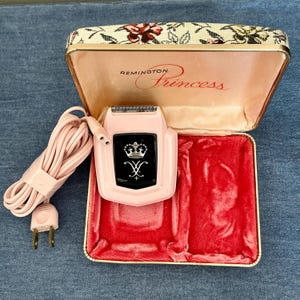 Pu&ograve; includere: Rasoio elettrico Remington Princess rosa vintage con un emblema in bianco e nero, nella sua custodia aperta a motivi floreali. La custodia ha un interno in velluto rosa. Sono visibili anche un cavo e una spina rosa.