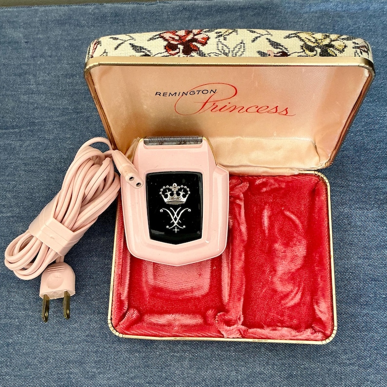 Pu&ograve; includere: Rasoio elettrico Remington Princess rosa vintage con un emblema in bianco e nero, nella sua custodia aperta a motivi floreali. La custodia ha un interno in velluto rosa. Sono visibili anche un cavo e una spina rosa.