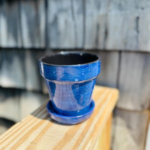 Może przedstawiać: Niebieska ceramiczna doniczka z pasującym spodkiem. Doniczka ma błyszczące wykończenie z nakrapianym wzorem i czarnym wnętrzem. Stoi na drewnianej powierzchni na tle zwietrzałych szarych desek.