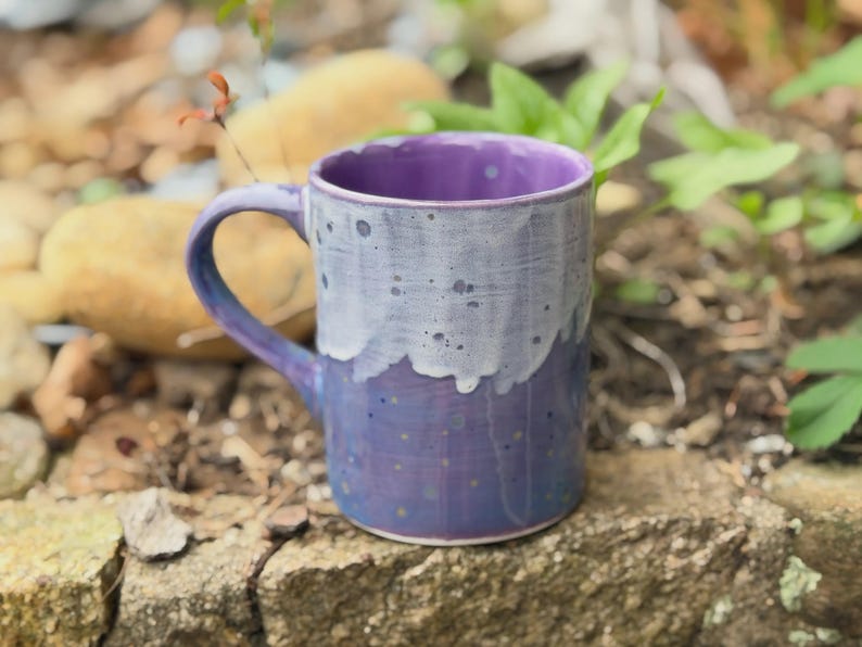 K&ouml;nnte beinhalten: Ein Keramikbecher mit zweifarbiger Glasur, bestehend aus einer tiefvioletten Basis und einer helleren, tropfenden wei&szlig;en Glasur im oberen Bereich. Die Oberfl&auml;che ist mit kleinen dunklen Sprenkeln versehen und das Innere ist einfarbig violett.