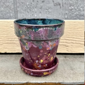 May include: A ceramic flower pot with a matching saucer. The pot is a deep burgundy color with a glossy finish and a speckled pattern of yellow, orange, and blue. The rim is a teal color.