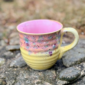 Puede incluir: Una taza de cerámica hecha a mano con asa y base amarillas e interior rosa. El exterior presenta un diseño moteado con detalles en naranja, verde y morado. La taza está sobre una superficie de piedra.