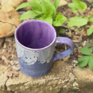 K&ouml;nnte beinhalten: Ein Keramikbecher mit leuchtend violettem Innenraum und einem Henkel. Die Au&szlig;enseite zeigt eine zweifarbige Glasur mit einer tiefblauen Basis, die in ein helleres, gesprenkeltes wei&szlig;es Tropfmuster am Rand &uuml;bergeht und eine Textur erzeugt.