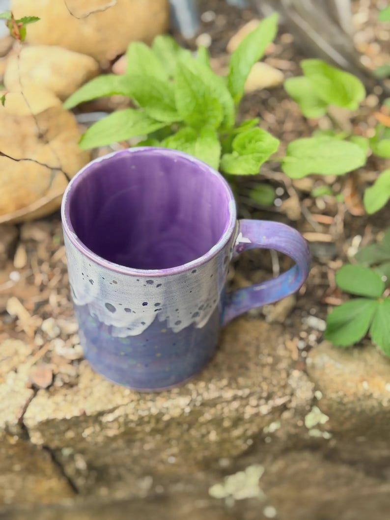 K&ouml;nnte beinhalten: Ein Keramikbecher mit leuchtend violettem Innenraum und einem Henkel. Die Au&szlig;enseite zeigt eine zweifarbige Glasur mit einer tiefblauen Basis, die in ein helleres, gesprenkeltes wei&szlig;es Tropfmuster am Rand &uuml;bergeht und eine Textur erzeugt.