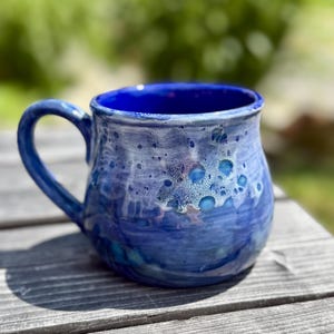 May include: A handmade ceramic mug with a blue glaze. The mug features a rounded body, a sturdy handle, and a deep blue interior. The exterior has a textured, speckled appearance with shades of blue and white.