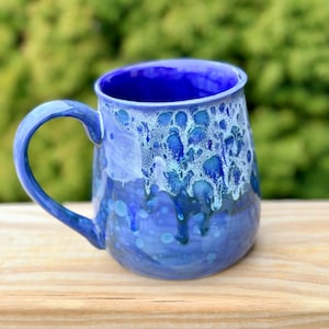 May include: A set of three ceramic mugs with a speckled, cream-colored glaze. Each mug features a different hand-painted design: a blue and green leaf, a yellow flower, and a red berry. The mugs have a simple, rounded shape.