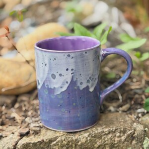K&ouml;nnte beinhalten: Ein Keramikbecher mit gl&auml;nzender Oberfl&auml;che, der eine tiefviolette Grundfarbe aufweist, die in ein helleres lavendelfarbenes und wei&szlig;es Tropfmuster am Rand &uuml;bergeht. Der Becher hat einen geschwungenen Henkel und einen glatten, zylindrischen K&ouml;rper.