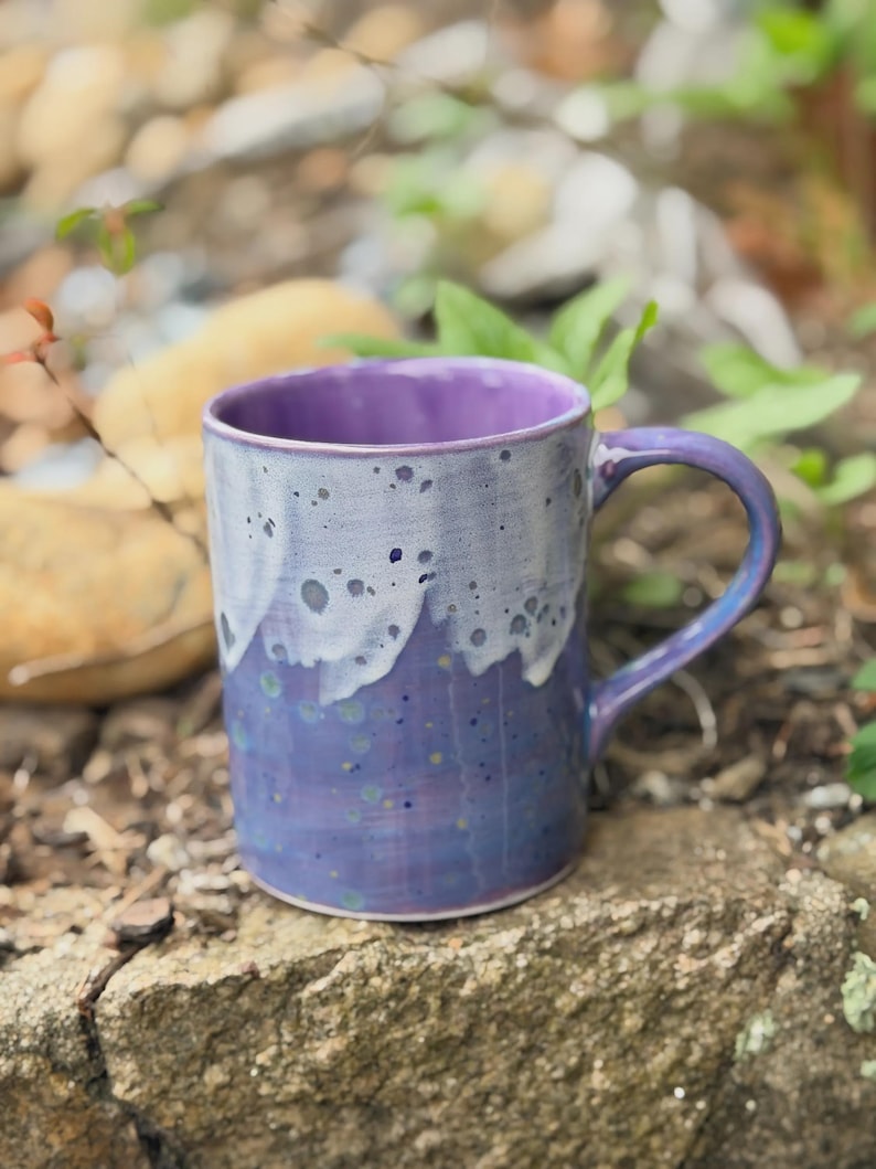 K&ouml;nnte beinhalten: Ein Keramikbecher mit gl&auml;nzender Oberfl&auml;che, der eine tiefviolette Grundfarbe aufweist, die in ein helleres lavendelfarbenes und wei&szlig;es Tropfmuster am Rand &uuml;bergeht. Der Becher hat einen geschwungenen Henkel und einen glatten, zylindrischen K&ouml;rper.
