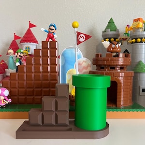 以下が含まれることがあります： 緑色のパイプ、茶色のレンガのプラットフォーム、キャラクターのフィギュアが特徴の、スーパーマリオブラザーズをテーマにしたディスプレイ。シーンには、城、旗、その他のビデオゲームの象徴的な要素が含まれています。