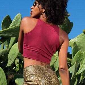 May include: A woman wearing a maroon tank top and gold sequined pants stands in front of a cactus.