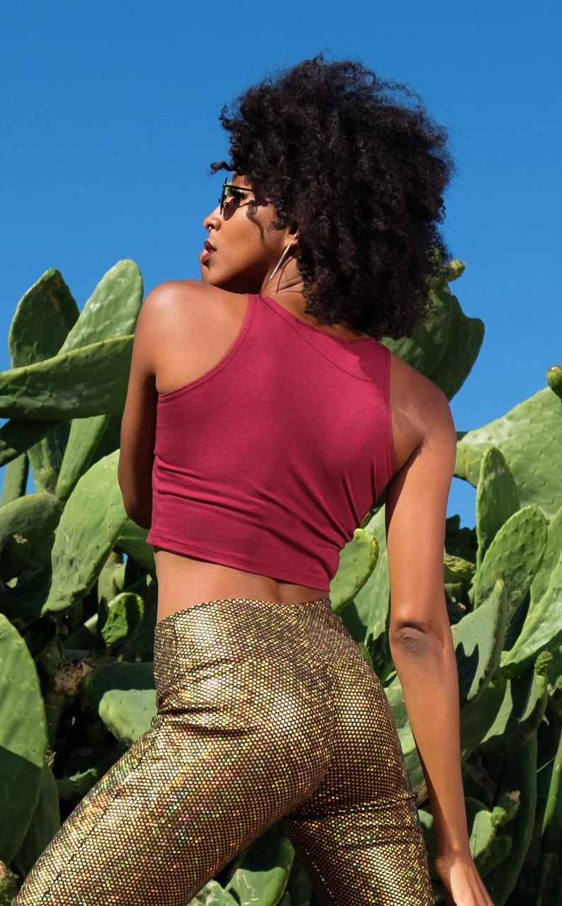 May include: A woman wearing a maroon tank top and gold sequined pants stands in front of a cactus.