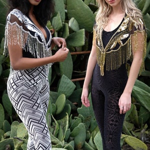 May include: Two women wearing black tops with sequined fringe shawls. One woman is wearing a black and white patterned pants and a black cowboy hat. The other woman is wearing black pants with a floral pattern. They are standing in front of a cactus.