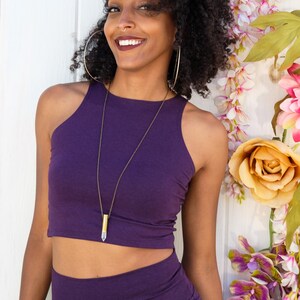 May include: A woman wearing a purple crop top and matching pants. She is wearing a gold necklace with a clear crystal pendant. She is smiling and looking at the camera.