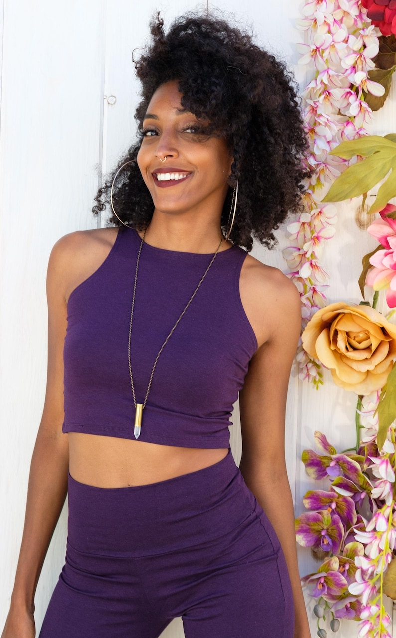 May include: A woman wearing a purple crop top and matching pants. She is wearing a gold necklace with a clear crystal pendant. She is smiling and looking at the camera.