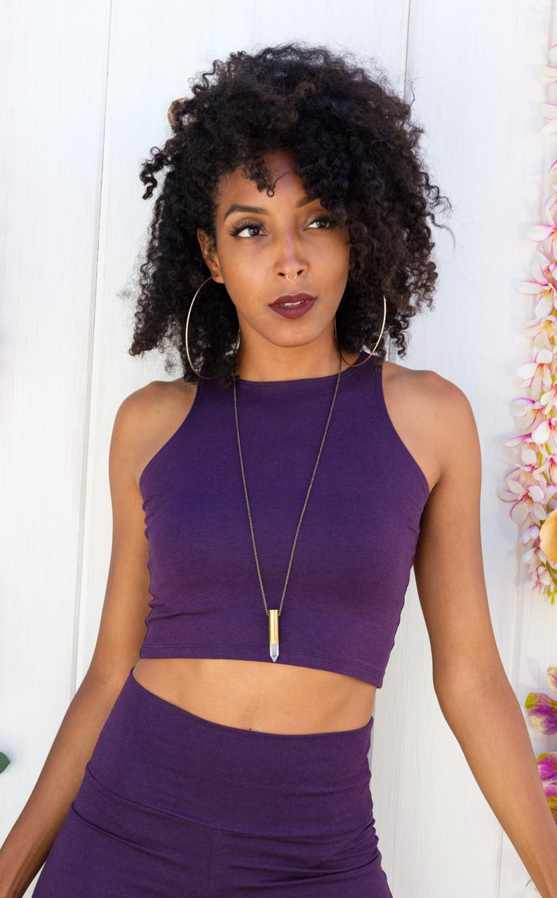 May include: A woman wearing a purple crop top and matching pants. She is wearing a gold necklace with a clear crystal pendant.