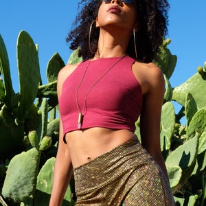 May include: A woman wearing a maroon crop top with a gold chain necklace and a crystal pendant. She is also wearing gold sparkly pants and black sunglasses. She is standing in front of a cactus.