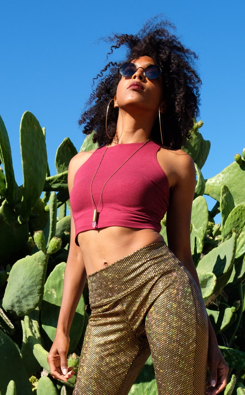 May include: A woman wearing a maroon crop top with a gold chain necklace and a crystal pendant. She is also wearing gold sparkly pants and black sunglasses. She is standing in front of a cactus.