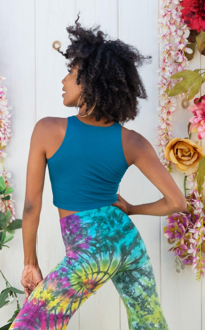 May include: A woman wearing a teal blue crop top and tie-dye pants. The pants have a swirl pattern in shades of blue, green, yellow, pink, and purple.