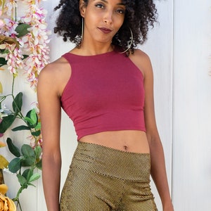 May include: A woman with curly dark hair is wearing a burgundy crop top and gold patterned pants. She is standing in front of a white wall with pink flowers.